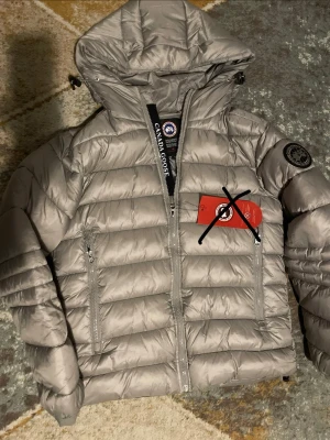 Ljusgrå dunjacka från Canada Goose - Snygg ljusgrå dunjacka från Canada Goose med huva och svarta detaljer. Jackan har två dragkedjeförsedda fickor framtill och en patch med logga på ärmen. Perfekt för kalla vinterdagar. Taggen ni ser på bilderna finns inte kvar! Därför jag lagt ett kryss på de!