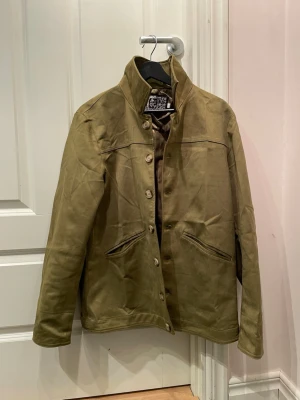 Olivgrön mockajacka med knappar - Snygg olivgrön mockajacka med ståkrage och knäppning framtill. Jackan har två snedställda fickor och en stilren, minimalistisk design. Perfekt för dig som vill ha en unik jacka med skön passform och modern look. Helt oanvänd. 