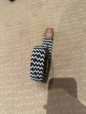 Randigt blåvitt flätat bälte 103 cm långt  - Snyggt flätat bälte i marinblått och vitt med randigt mönster. Bältet har detaljer i brunt skinn vid spännet och änden, samt ett klassiskt metallspänne. Perfekt accessoar för att lyfta din outfit. Kontakta vid minsta intresse!