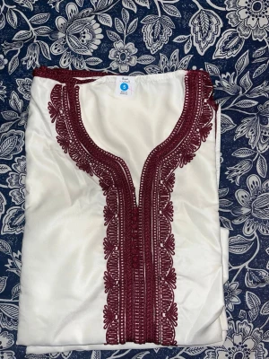 Vit kaftan med vinröd brodyr - Snygg vit kaftan med detaljerad vinröd brodyr längs halsringningen och framtill. Plagget har en klassisk V-ringning och är tillverkat i ett glansigt, mjukt tyg som ger en lyxig känsla. Perfekt för dig som vill sticka ut med stilrena detaljer.