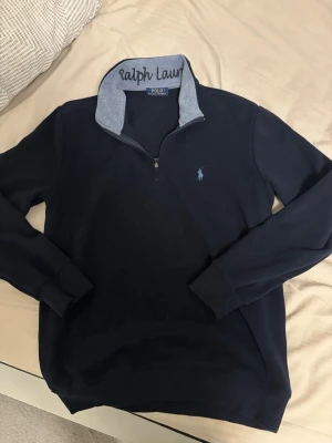 Mörkblå half-zip tröja Polo Ralph Lauren - Mörkblå långärmad tröja från Polo Ralph Lauren med half-zip och broderad logga på bröstet. Insidan av kragen är ljusblå med Ralph Lauren-broderi. Tillverkad i mjukt bomullsmaterial och har ribbade muddar vid ärmslut och nederkant. OBS: storlek S inte M
