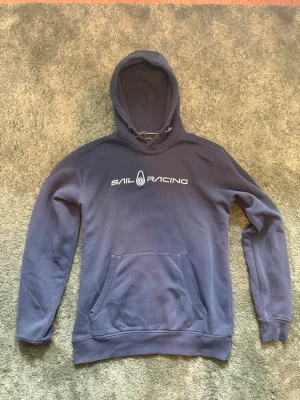 Sail Racing hoodie blå - Mörkblå hoodie från Sail Racing med vit logga och text på bröstet. Klassisk ficka på magen framtill, ribbade muddar vid ärmarna och nederkant samt justerbar huva med snören. Hoodien är i sjukt bra skick och är knappt använd🤩Perfekt för de kallare dagarna❄️ Plagget fraktas samma dag det är köpt❤️‍🩹 Pris kan diskuteras vid en snabb affär💨