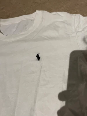 Vit t-shirt med broderad logga - Vit t-shirt med rund hals och korta ärmar. På bröstet finns en liten broderad logga i svart. Enkel och stilren design som passar till det mesta. Tillverkad i mjukt material för skön känsla.