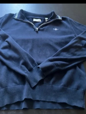 Mörkblå stickad tröja från Gant - Mörkblå stickad tröja från Gant med halv dragkedja och ribbade muddar. Tröjan har en liten broderad logga på bröstet och randig detalj vid kragen. Perfekt för en stilren och avslappnad look.