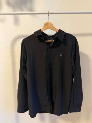Svart skjorta från polo - Stilren svart skjorta med klassisk krage och knäppning framtill. Skjortan har lång ärm och en broderad logga på bröstet i vitt. Perfekt för dig som gillar en enkel men snygg look.
