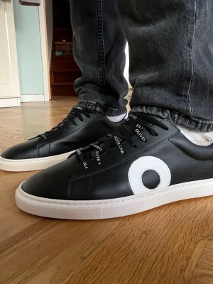 Svarta sneakers från Oliver Cabell  - Snygga svarta sneakers med vit sula och en stor vit cirkeldetalj på sidan. Skorna har svarta skosnören och en låg profil som passar till många olika stilar.