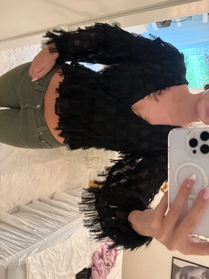 Svart croppad blus med fransar - Säljer en svart croppad blus med v-ringning och långa ärmar. Blusen har unika fransdetaljer och ett transparent, luftigt material som ger en cool och trendig look. Perfekt för dig som vill sticka ut lite extra.