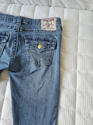 Blå bootcut jeans från True Religion - Snygga blå bootcut jeans från True Religion med klassiska kontrastsömmar och ficklock bak med guldfärgad knapp. Jeansen har en avslappnad passform och är tillverkade i mjukt denimtyg. Perfekta för en trendig och avslappnad stil. Den har gått sönder en gång då vi lagat den och nu syns det knappt, som ni ser på bilderna 🤗🤗