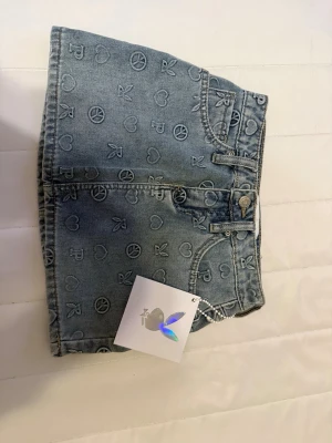 Playboy  jeanskjol med peace- och hjärtmönster - Säljer en helt ny blå jeanskjol från playboy med all-over tryck av peace-symboler, hjärtan och bokstaven P. Kjolen har klassisk femficksdesign, bälteshällor och knappstängning framtill. Perfekt för dig som gillar en lekfull och trendig stil. Passar bäst dig som använder XXS-XS, kika ändå gärna dina mått