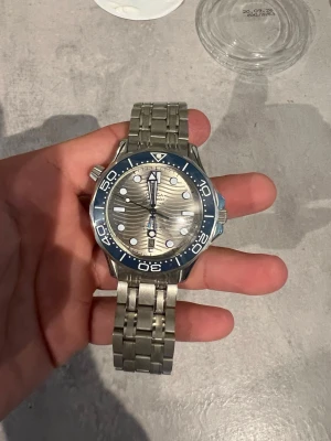 Seamaster blå/silver klocka - Säljer en  Seamaster klocka med blå vridring och silverfärgad urtavla med vågmönster. Klockan har en robust länk i rostfritt stål och tydliga indexmarkeringar. Perfekt för dig som gillar klassisk och sportig stil.