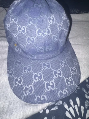 Blå keps med GG-mönster från Gucci - Snygg blå keps från Gucci med det klassiska GG-monogrammet över hela kepsen. Kepsen har böjd skärm och är tillverkad i ett tåligt tygmaterial. Insidan är ljus och har etikett med Gucci-logga. Perfekt för dig som vill sticka ut med en lyxig accessoar.