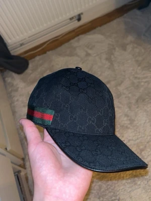 Svart Gucci keps med GG-mönster - Svart keps från Gucci med klassiskt GG-monogram över hela ytan. Kepsen har en böjd skärm och detaljeras med en grön och röd rand på sidan. Materialet ser ut att vara canvas med läderdetaljer vid skärmen. Kommer i storlek L/59 och är justerbar. 