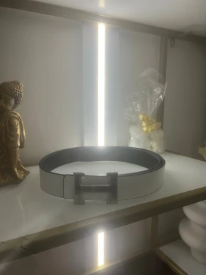 Vitt bälte H med silverspänn 120cm - Stilrent vitt bälte med ett stort, fyrkantigt metallspänne. Bältet har en slät yta och klassisk design som passar till många olika outfits. Perfekt accessoar för att lyfta din look.