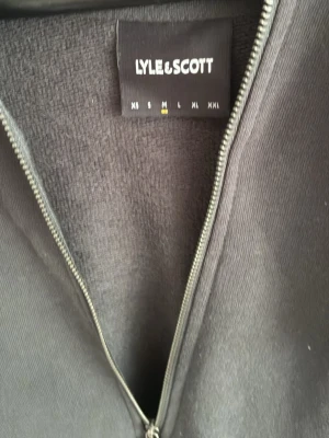 Mörkgrå hoodie från Lyle & Scott - Säljer en stilren mörkgrå hoodie från Lyle & Scott i storlek M. Hoodien har dragkedja framtill, huva och ribbade muddar vid ärmslut och nederkant. Tillverkad i mjukt bomullsmaterial som känns skönt mot huden. Perfekt för en avslappnad look.