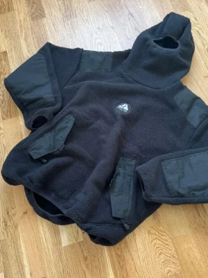 Nike ACG Ninja Fleece - Säljer en svart teddyjacka från Nike ACG med hög krage och huva. Jackan har förstärkta partier på armbågar och axlar, samt två stora fickor med dragkedja framtill. Tillverkad i mjukt teddy-material, perfekt för kalla dagar.