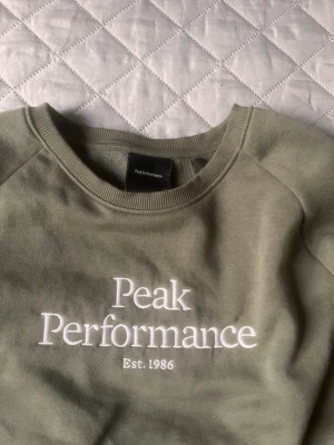 Grön sweatshirt Peak Performance - Stilren grön sweatshirt från Peak Performance med vit broderad logga och text 'Est. 1986' framtill. Tröjan har rund halsringning, långa ärmar och ribbade muddar. Perfekt för en avslappnad och sportig look.