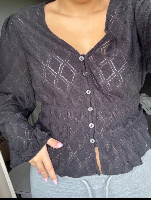Svart blus med knappar  - Snygg svart blus med v-ringning och dekorativa knappar framtill från Lindex. Blusen har ett strukturerat tyg med diskreta rutmönster och markerad midja. Långärmad modell. Storlek S