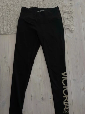 Victoria Sport leggings - Snygga svarta leggings med stor guldfärgad text 'VICTORIA' längs ena benet. Tight passform och hög midja, perfekta till både träning och chill. Materialet är stretchigt och mjukt för maximal komfort.