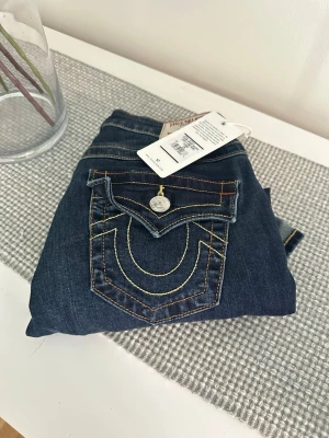 Mörkblå True Religion jeans - Snygga mörkblå jeans från True Religion med ikoniska kontrastsömmar och stora fickor bak med dekorativa knappar. Klassisk femficksmodell i mjukt denimtyg. Perfekta för dig som gillar streetwear och vill sticka ut med detaljer.