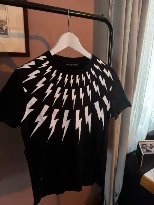 Svart t-shirt med blixtar Neil Barrett - Säljer en svart t-shirt från Neil Barrett med vita blixtmönster runt halsen och axlarna. T-shirten har rund halsringning och korta ärmar. Tillverkad i mjuk bomull för en bekväm känsla och cool look.