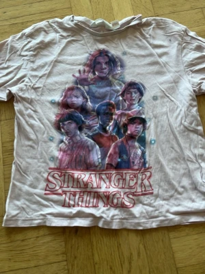 Stranger Things t-shirt från H&M - Cool vit Stranger Things t-shirt från H&M med färgglatt tryck av karaktärerna från serien och stor logga framtill. Perfekt för dig som älskar Netflix och vill ha en unik tee med snygg design. Rund hals och korta ärmar.