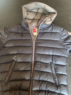 Grå dunjacka med huva från Colmar - Snygg grå dunjacka från Colmar med huva och dragkedja framtill. Jackan har två snedställda fickor med dragkedja och quiltad design. Perfekt för kyliga dagar och riktigt lätt att bära. Klassisk logga på ärmen, ett pytte litet håll på första bilden men jag tror inte de är några bekymmer , jag har knappt använt jackan och den är i jätte bra skick .
