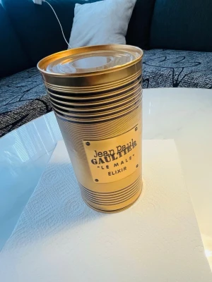Jean Paul Gaultier Le Male Elixir - Jean Paul Gaultier Le Male Elixir parfym i en lyxig guldfärgad metallförpackning. Flaskan har en unik design formad som en manlig torso i guldtoner och står på en röd sammetsliknande bas. Perfekt för dig som gillar exklusiva och iögonfallande dofter.