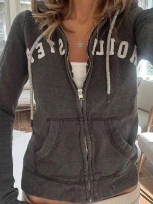 Mörkgrå Hollister hoodie med dragkedja - Säljer en mörkgrå hoodie från Hollister med vit text över bröstet. Hoodien har dragkedja framtill, två stora fickor och justerbar huva med snören. Perfekt för en chill och avslappnad stil.