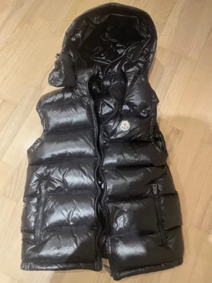 Svart dunväst med avtagbar huva - Snygg svart dunväst från Moncler med glansig finish och avtagbar huva. Västen har två dragkedjeförsedda fickor framtill och klassisk Moncler-logga på bröstet. Perfekt för lager-på-lager och håller dig varm under kyliga dagar.