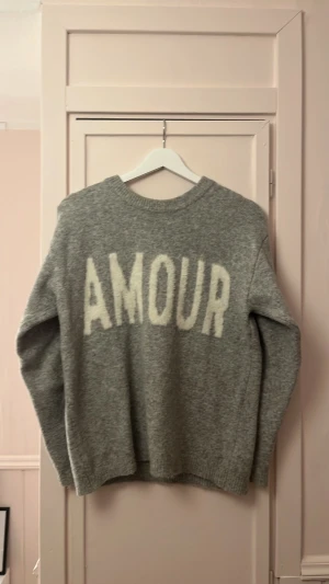 Grå stickad tröja med text - Mysig grå stickad tröja med rund hals och långa ärmar. På ryggen står det 'AMOUR' i stora vita bokstäver. Från Kappahl. Lite smink märken runt kragen (se bild 3)