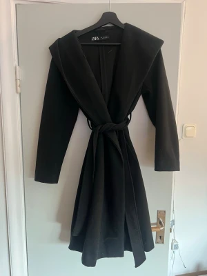Svart kappa med huva från Zara - Stilren svart kappa från Zara med stor huva och knytskärp i midjan. Kappan har lång ärm och en klassisk, något utsvängd passform. Perfekt för dig som vill ha en enkel men trendig look. Materialet ser ut att vara mjukt och lite tjockare, perfekt för kyligare dagar.