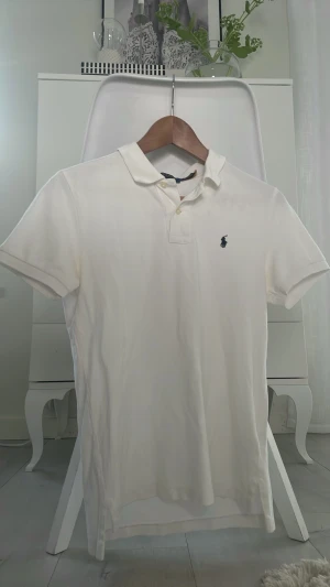 Vit pikétröja från Polo Ralph Lauren - Klassisk vit pikétröja från Polo Ralph Lauren i slim fit-modell. Tröjan har korta ärmar, krage och knappar framtill samt den ikoniska broderade loggan på bröstet. Perfekt för en stilren och fräsch look.