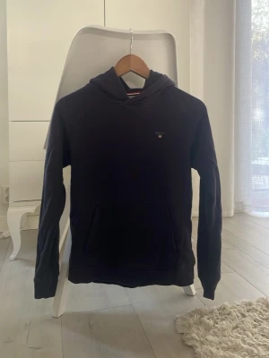 Mörkblå hoodie från GANT - Snygg mörkblå hoodie från GANT med klassisk huva och känguruficka framtill. Diskret GANT-logga på bröstet och mjukt material som passar perfekt till vardagslooken. Passar dig som gillar stilrena och bekväma plagg.