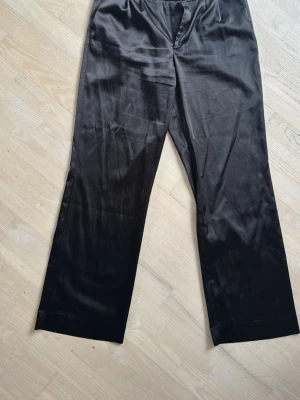 Sanne Alexandra Svarta vida kostymbyxor i satin - Snygga svarta kostymbyxor från Marc Cain i glansigt satinmaterial. Byxorna har hög midja, vida ben och dubbla knappar i midjan för en stilren look. Perfekta för dig som vill ha en dressad och trendig stil.