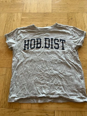 Grå t-shirt med tryck HOB.DIST - Säljer en grå t-shirt med svart texten HOB.DIST på bröstet. T-shirten har rund hals och korta ärmar. Materialet är mjuk bomullsmix som känns skön mot huden. Perfekt till jeans eller shorts för en avslappnad look.