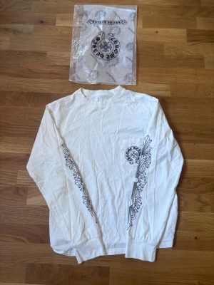 Vit långärmad Chrome Hearts tröja - Säljer en vit långärmad tröja från Chrome Hearts med svart tryck på bröstet och stort motiv på ryggen. Ärmarna har detaljerade svarta mönster längs sidorna. Tröjan har rund hals och ribbade muddar. Perfekt för dig som gillar streetwear och unika prints.