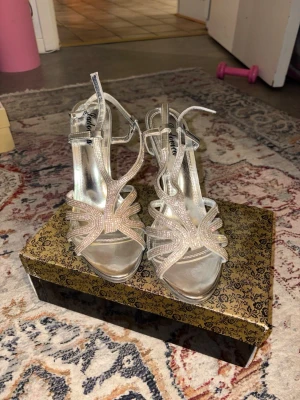 Silvriga strassandaler med hög klack - Säljer ett par glittriga silvriga sandaler med öppen tå och smala remmar dekorerade med strass. Skorna har hög stilettklack och justerbara ankelband. Perfekta för dig som vill sticka ut och glänsa lite extra. Materialet är syntet med metallicfinish.