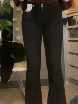 Svarta bootcut jeans med dubbla knappar - Säljer ett par svarta bootcut jeans med medelhög midja och dubbla knappar i linningen. Jeansen har klassisk femficksdesign och är tillverkade i stretchigt denimtyg för en bekväm passform. Perfekta för dig som gillar en tidlös men trendig look.