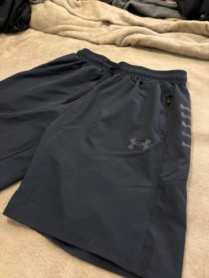 Mörkblå träningsshorts från Under Armour - Snygga mörkblå träningsshorts från Under Armour med elastisk midja och dragsko. Shortsen har dragkedjeficka och tryckt logga samt texten 'ARMOUR' längs ena benet. Perfekta för gymmet eller löprundan.