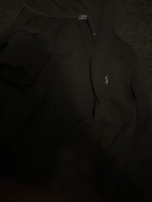 Svart hoodie med liten logga - Svart hoodie med huva och en liten broderad logga på bröstet. Klassisk modell med känguruficka framtill och ribbade muddar vid ärmslut och nederkant. Perfekt för en avslappnad stil.