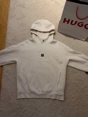 Vit hoodie från Hugo boss  - Snygg vit hoodie med huva och broderad logga på bröstet. Hoodien har känguruficka och ribbade muddar vid ärmslut och nederkant. Perfekt för en avslappnad och trendig stil.