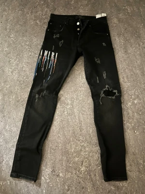 Svarta distressed jeans från Amiri - Säljer ett par svarta skinny jeans från Amiri med slitningar och färgstänk i blått, vitt och rött på vänster lår. Jeansen har knappgylf, fem fickor och coola detaljer som ger en edgy vibe. Perfekta för dig som gillar streetwear och unik design.
