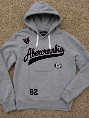 Grå hoodie från Abercrombie - Snygg grå hoodie från Abercrombie med broderad logga i vinrött och svart framtill. Har huva med vita snören, känguruficka och detaljer som siffran 92 och patchar. Perfekt för en avslappnad och sportig stil.