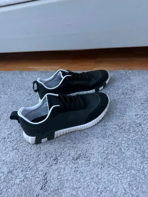 Svarta sneakers med vit sula - Snygga svarta sneakers med vit, räfflad sula och svarta skosnören. Skorna har vita detaljer runt öppningen och en modern, sportig look. Materialet ser ut att vara en mix av syntet och textil. Perfekta för dig som gillar stilrena och bekväma skor.