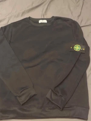 Svart sweatshirt från Stone Island - En svart sweatshirt från Stone Island med klassisk rund hals och den ikoniska logotypen på vänster ärm. Tröjan har ribbade muddar vid ärmslut och nederkant. Perfekt för en avslappnad och stilren look.