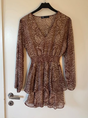Brun mönstrad kortklänning från Zara - Supersnygg kortklänning från Zara i brun och beige ton med ett coolt ormliknande mönster. Klänningen har v-ringning, långa ärmar och volangdetaljer i kjolen. Perfekt för dig som vill sticka ut lite extra!