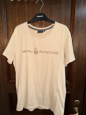 Beige t-shirt från Sail Racing - Snygg beige t-shirt från Sail Racing med tryckt logga på bröstet. Klassisk rund halsringning och korta ärmar. Tillverkad i mjuk bomull som känns skön mot huden. Perfekt för en avslappnad och stilren look.