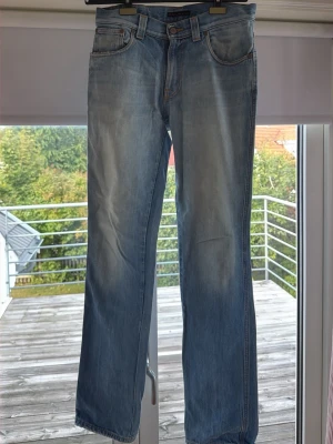 Nudie jeans Ljusblå raka  - Säljer ett par ljusblå jeans med rak passform och klassisk femficksdesign. Jeansen har subtila slitningar framtill och kontrastsömmar. Materialet är denim i bomull och bakfickorna har en diskret broderad detalj.