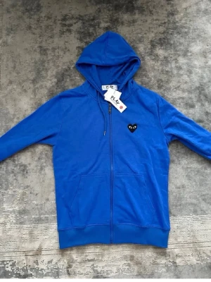 Blå hoodie med dragkedja och hjärtdetalj - Snygg blå hoodie med dragkedja framtill och huva. På bröstet sitter en svart hjärtdetalj med ögon, vilket ger en unik touch. Hoodien har två fickor framtill och ribbade muddar vid ärmar och nederkant. Perfekt för en chill och avslappnad stil.