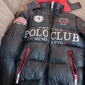 Svart pufferjacka från Santoz Polo Club - Säljer en svart pufferjacka från Santoz Polo Club med coola tryck och broderier. Jackan har en dragkedja och röda detaljer på fickorna. Insidan är röd med ett stort emblem. Perfekt för vintern och hösten!